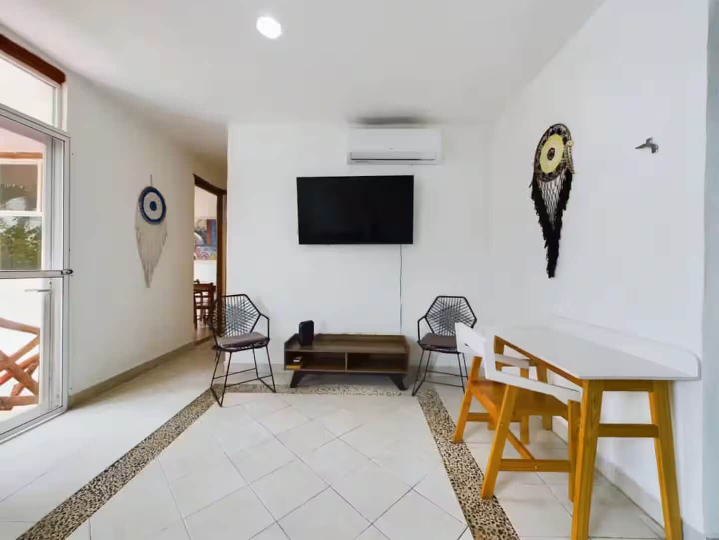 A modern living room with a flat-screen TV mounted on the wall, two chairs, a wooden dining table, and a decorative wall piece.