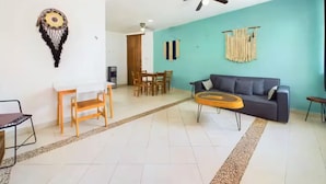 Ein Wohnzimmer mit einer türkisfarbenen Akzentwand, einem dunkelgrauen Sofa, einem Holzesstisch mit Stühlen, einem Deckenventilator und einem Wandbehang aus Makramee.