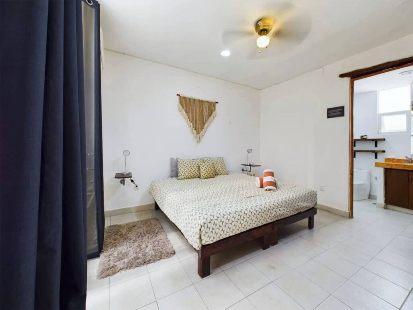 Un dormitorio con cama, ventilador de techo, una alfombra pequeña y un baño visible a través de una puerta abierta.