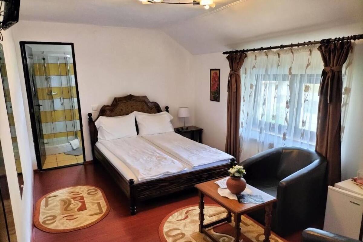 A hotel room with a bed, a chair, a desk, a small table, a nightstand, a mirror, a shower, and a window with curtains.