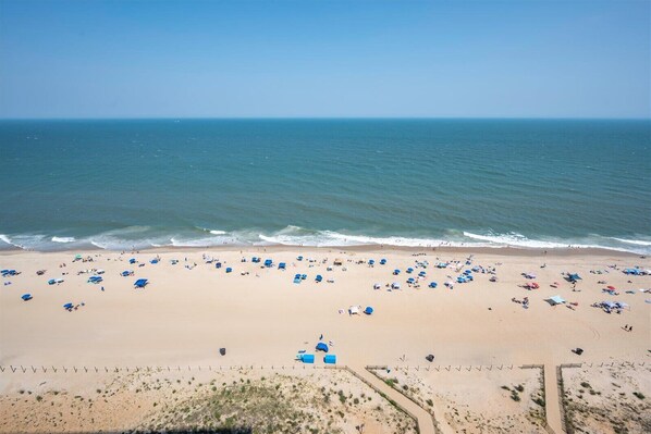 Uma praia com vários guarda-sóis, pessoas, céu claro e mar calmo.