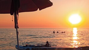 Un coucher de soleil sur une plage, avec un bateau et des personnes dans l’eau.