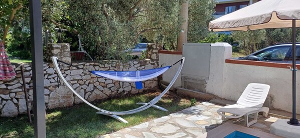 A hammock hanging between stone walls, a white plastic chair, and a poolside area with a stone pathway.