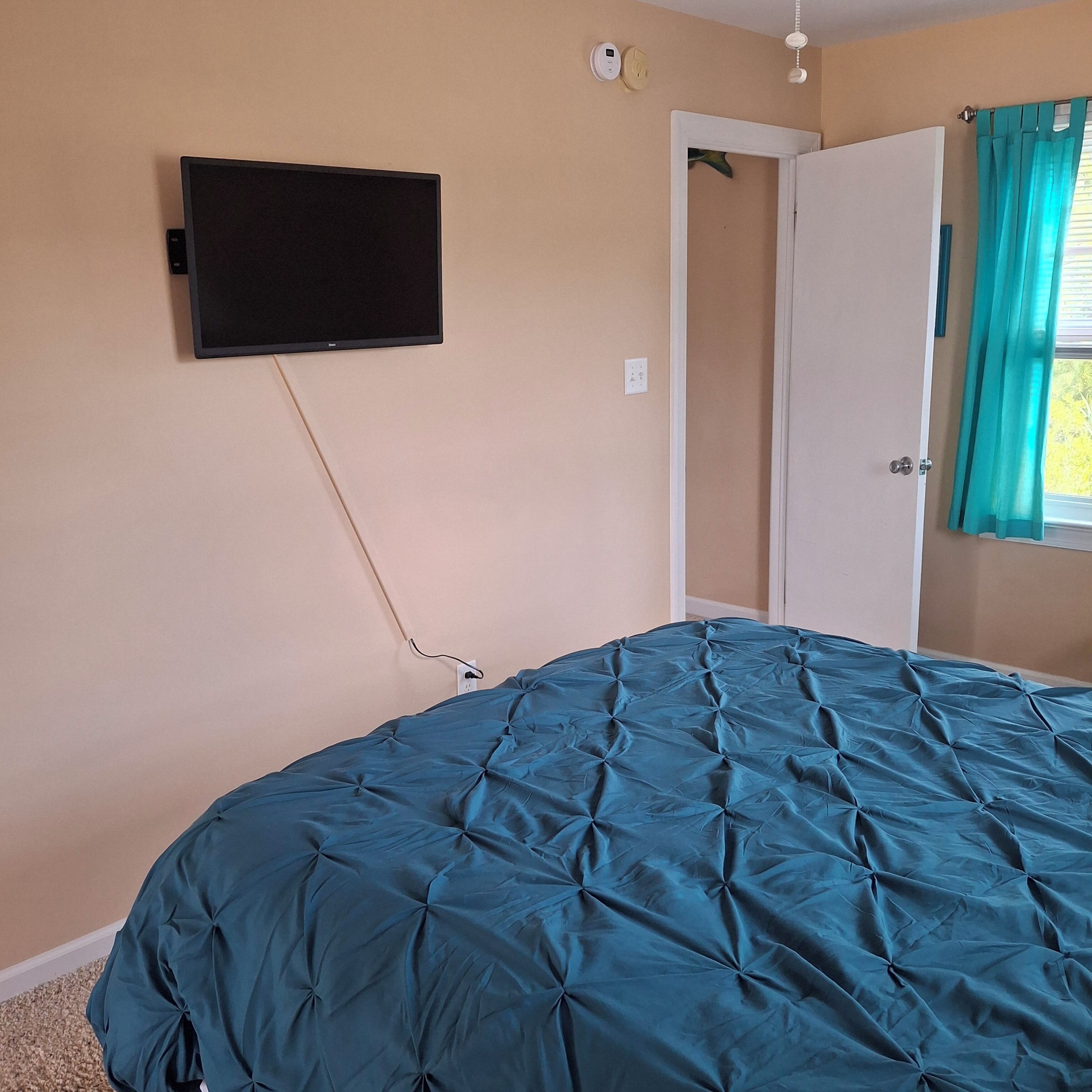 Ein Schlafzimmer mit Bett, Wandfernseher, Tür und einem Vorhangfenster.
