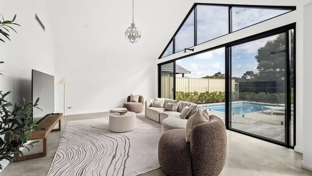 A modern living room with a sectional sofa, a round stone chair, a glass-enclosed patio, and a large window overlooking a pool. - Ember & Aqua—Luxury Barnhouse on the Golf Course (Dunsborough)