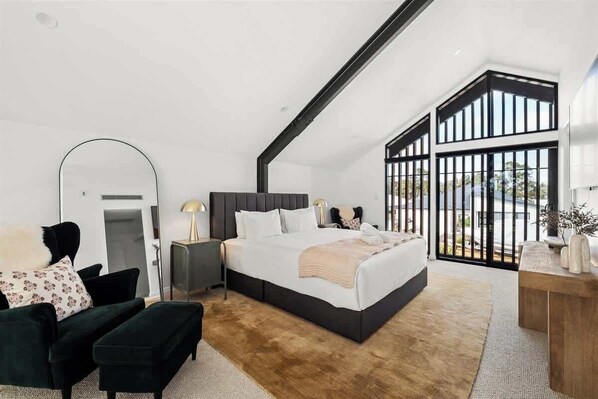A modern bedroom with a large bed, a dark headboard, a wooden side table, and a view through a large window.