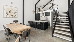 A modern kitchen with a central island, a dining area with a wooden table and chairs, and a black staircase. - Ember & Aqua—Luxury Barnhouse on the Golf Course (Dunsborough)