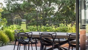 An outdoor patio with a wooden table and black chairs, surrounded by greenery and a black fence. - Ember & Aqua—Luxury Barnhouse on the Golf Course (Dunsborough)