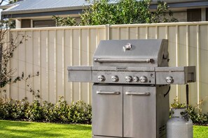 A stainless steel outdoor grill with four control knobs and a gas tank, set against a residential backdrop with a fence and greenery.