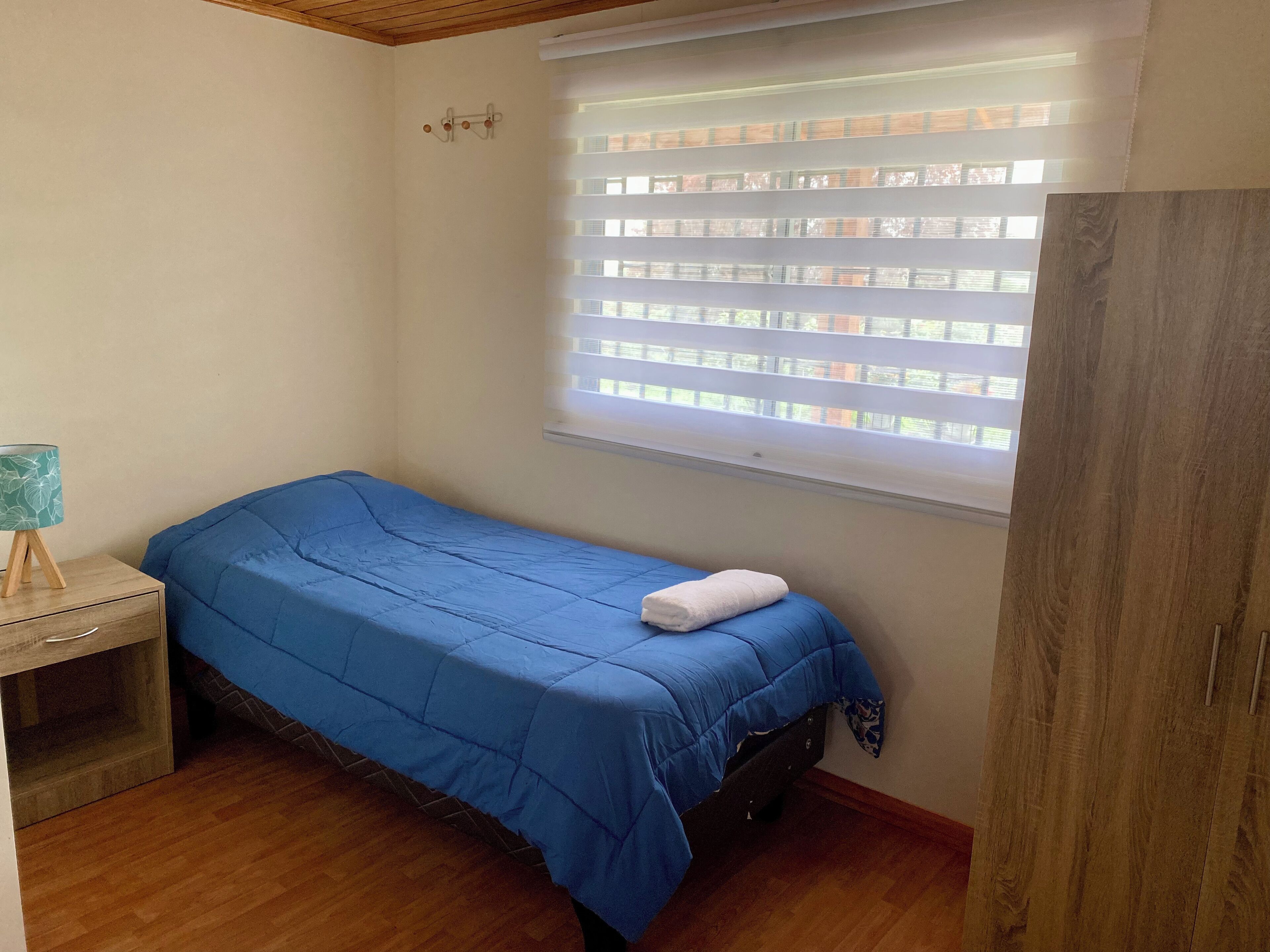 Een kleine kamer met een enkel bed, een houten vloer, een nachtkastje met een lamp en een raam met zonwering.