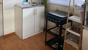 Eine kleine Küche mit Kühlschrank, Spüle und einem kleinen Herd. Ein Korkwandbrett zeigt Notizen und ein Spielzeug-Lkw, und ein Fenster mit Jalousien.