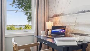 Un escritorio con una laptop, una lámpara y una taza, situado cerca de una ventana con vista a un lago y árboles.