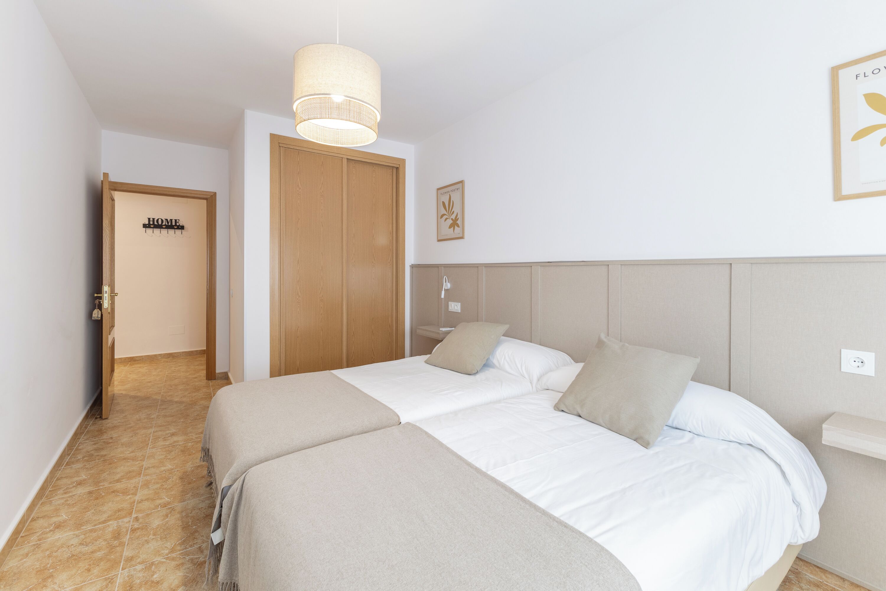 Un dormitorio con dos camas, un armario de madera, una lámpara colgante y un cuadro en la pared.