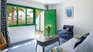 Une pièce avec une porte verte, une fenêtre donnant sur des palmiers, une chaise bleue et un lit avec des coussins à motifs.