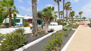 A landscaped area with palm trees, cacti, and a white building with green trim. - Vacation 'Jardin Del Sol 12' with Mountain View, Shared Pool and Wi-Fi (Playa Blanca)