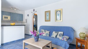 A modern living room with a blue sofa, a wooden coffee table, and a kitchen area with a microwave. - Vacation 'Jardin Del Sol 12' with Mountain View, Shared Pool and Wi-Fi (Playa Blanca)