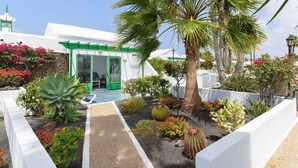 A white building with a green door, surrounded by various plants and a palm tree. - Vacation 'Jardin Del Sol 12' with Mountain View, Shared Pool and Wi-Fi (Playa Blanca)