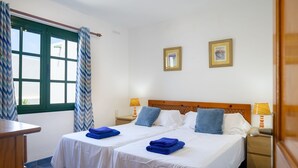 A double bed with two pillows, a nightstand with a lamp, and a window with curtains. - Vacation 'Jardin Del Sol 12' with Mountain View, Shared Pool and Wi-Fi (Playa Blanca)
