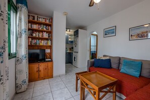 Un salon avec un canapé, une table basse en bois, une bibliothèque et, au fond, une cuisine.