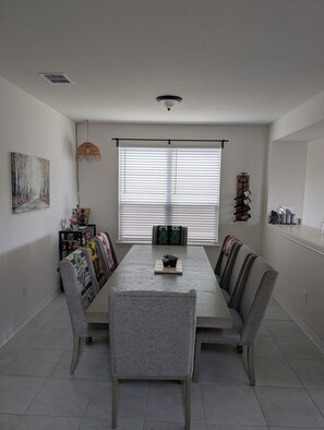 A dining area with a long table, grey upholstered chairs, a hanging light fixture, and a window with blinds. - Light and Airy 4-bedroom home in New Braunfels (New Braunfels)