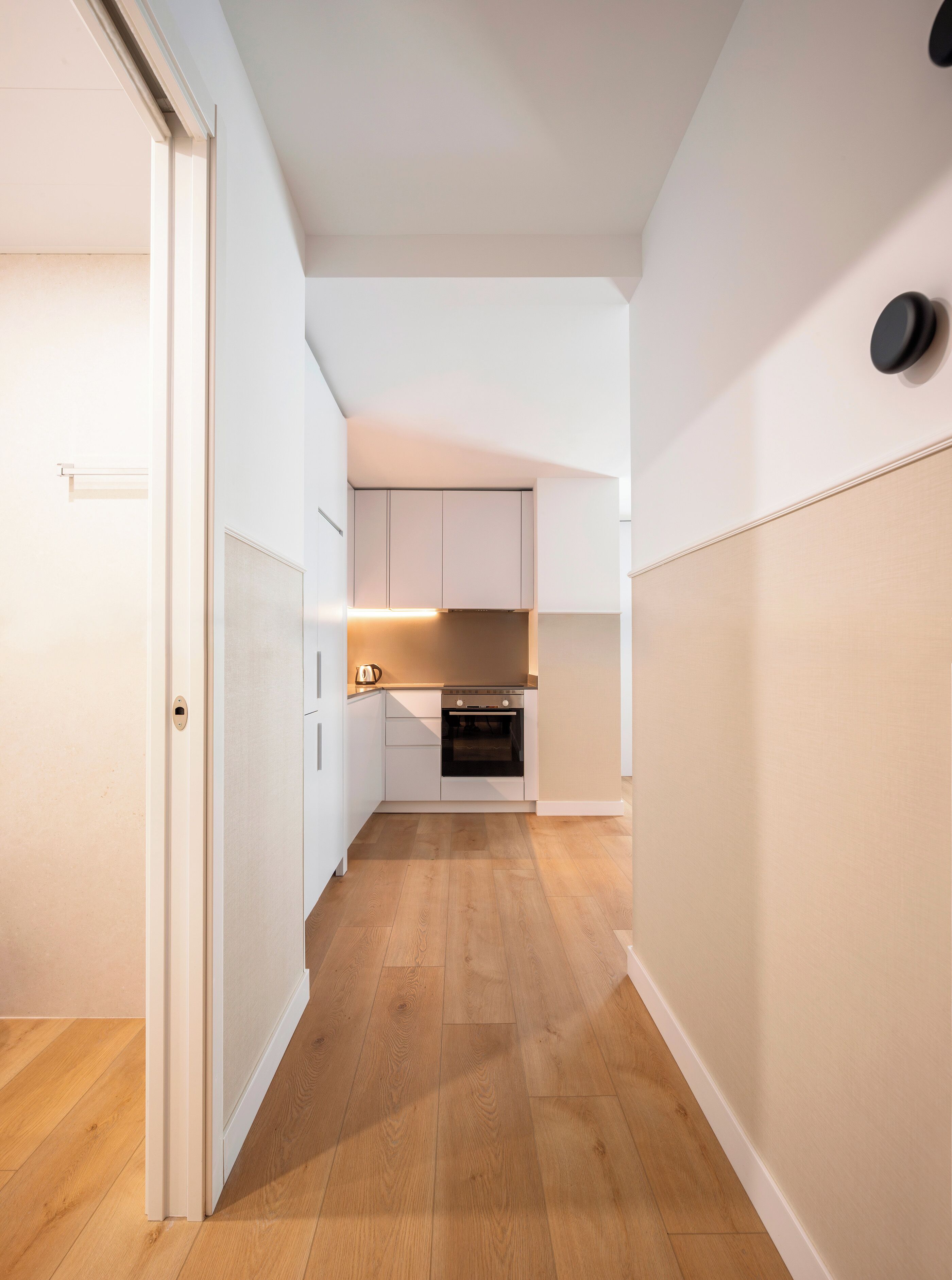 A narrow hallway with wooden flooring leading to a kitchen area with white cabinets and a black oven.
