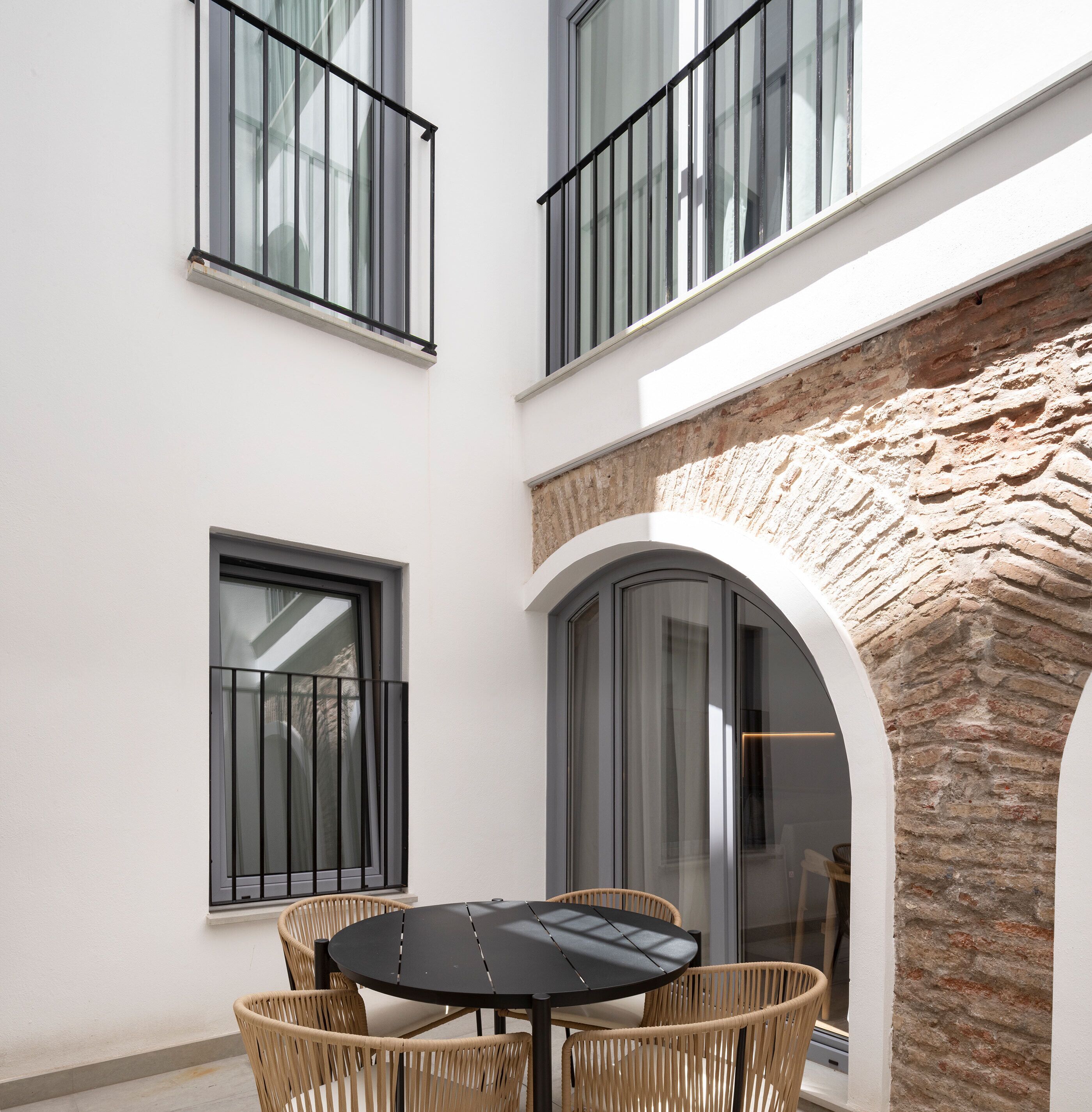 A modern outdoor patio with a round table and wicker chairs, a stone wall, and large windows with black railings.