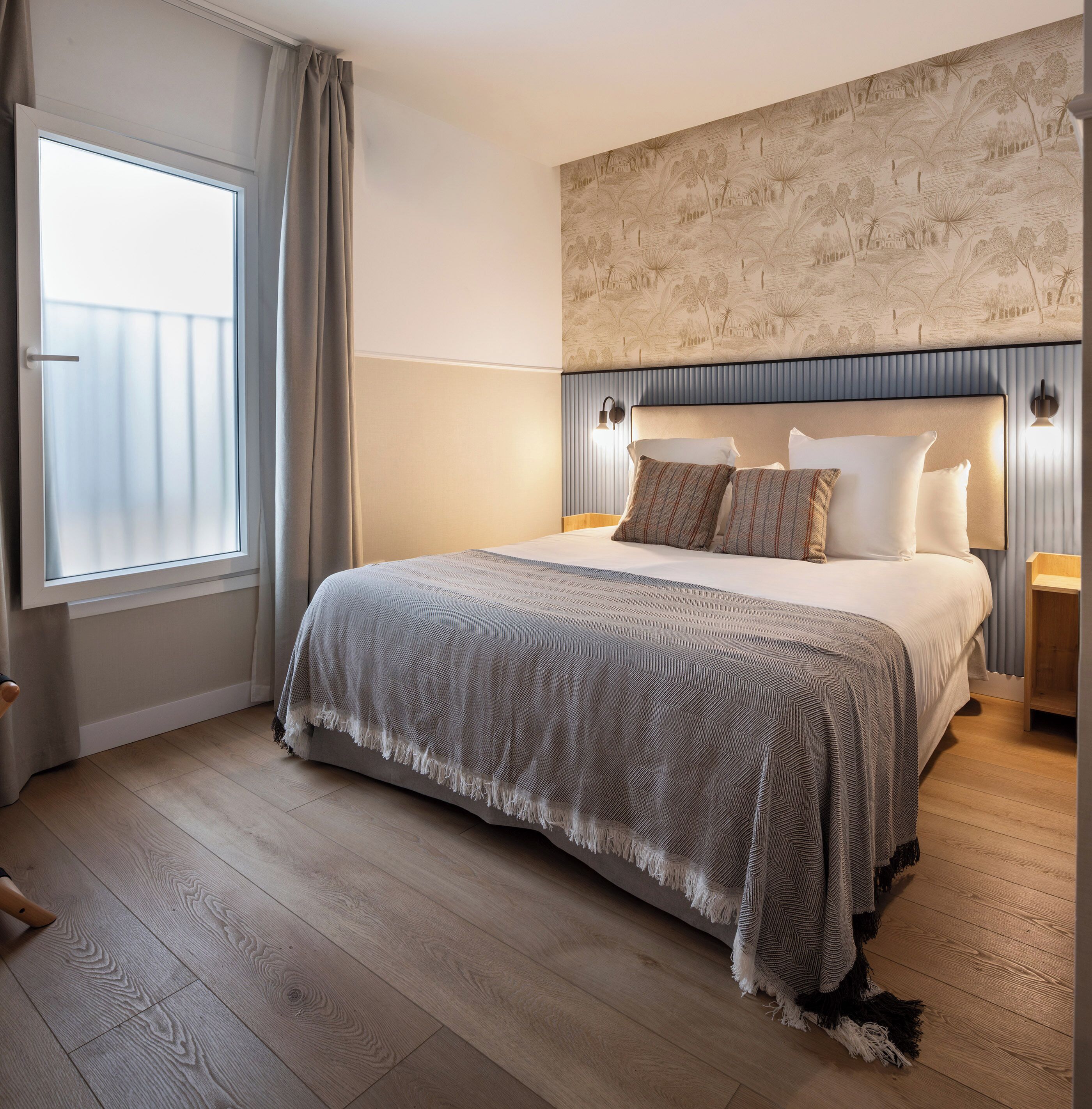 A hotel room with a bed, a large window, a wall with a mural, and a wooden floor.