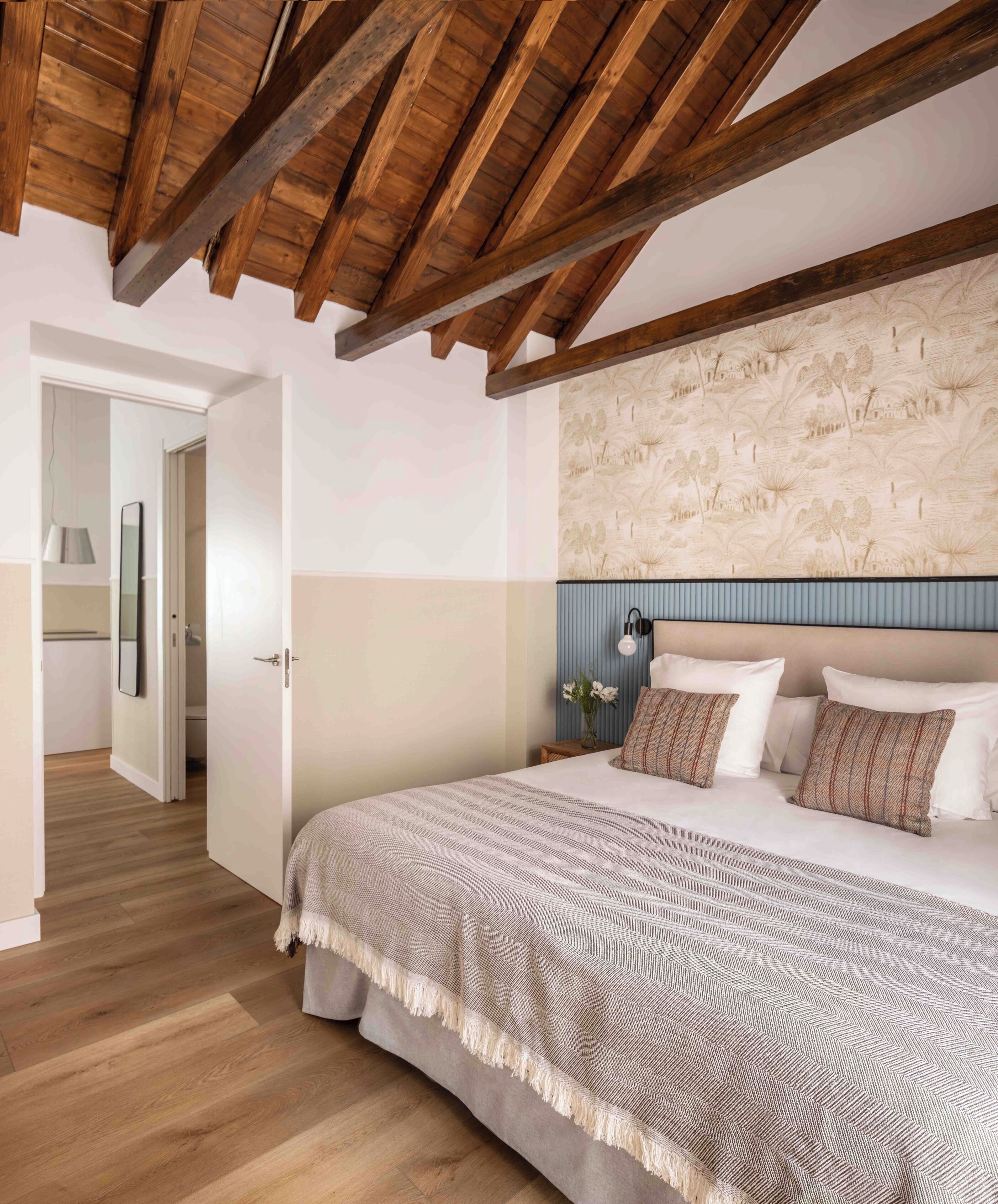 Un dormitorio con techo de madera, una cama con colcha estampada y una pared con papel tapiz botánico.