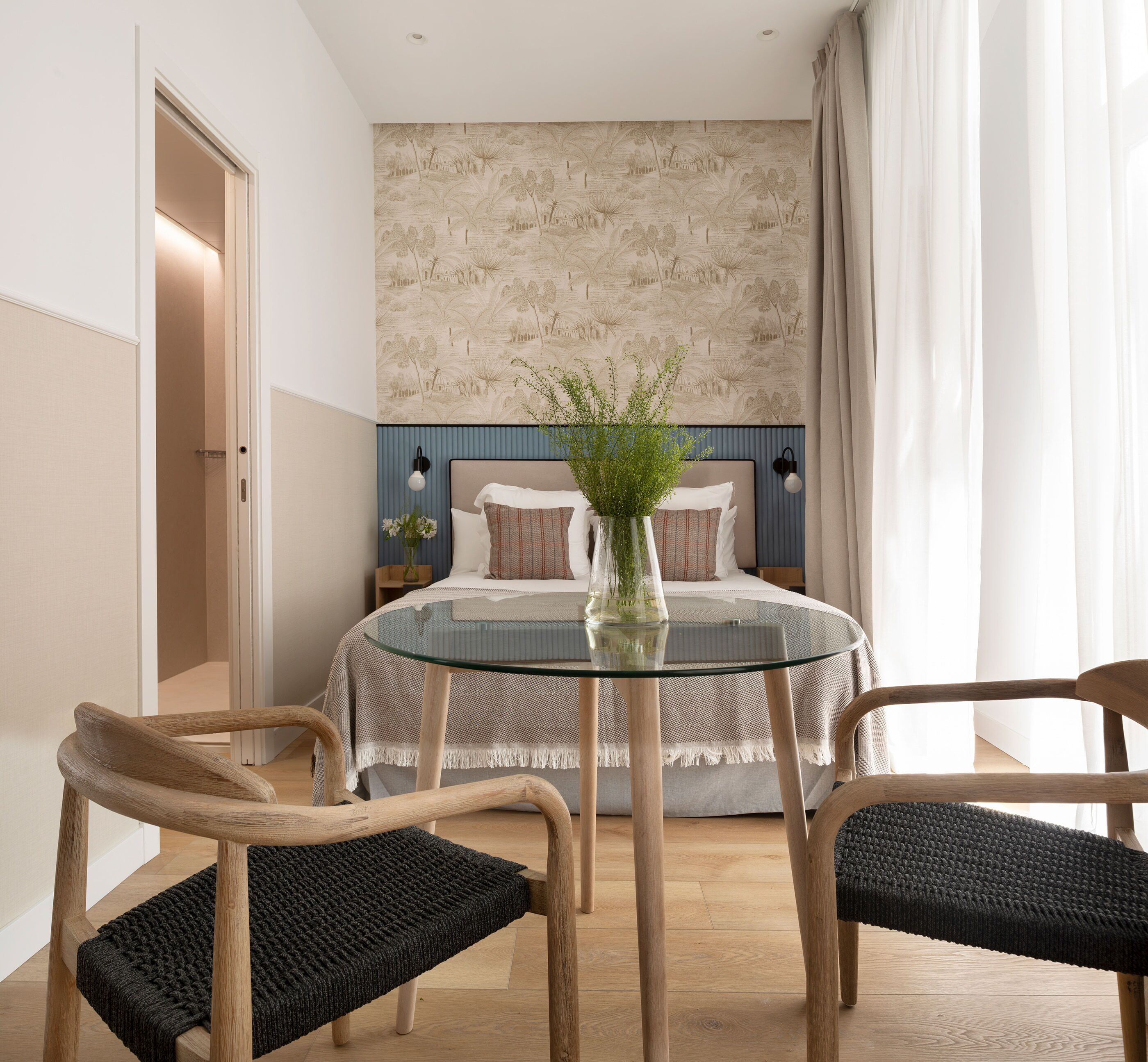 Ein modernes Schlafzimmer mit einem Bett, zwei Stühlen, einem Glastisch und einer Zierpflanze.