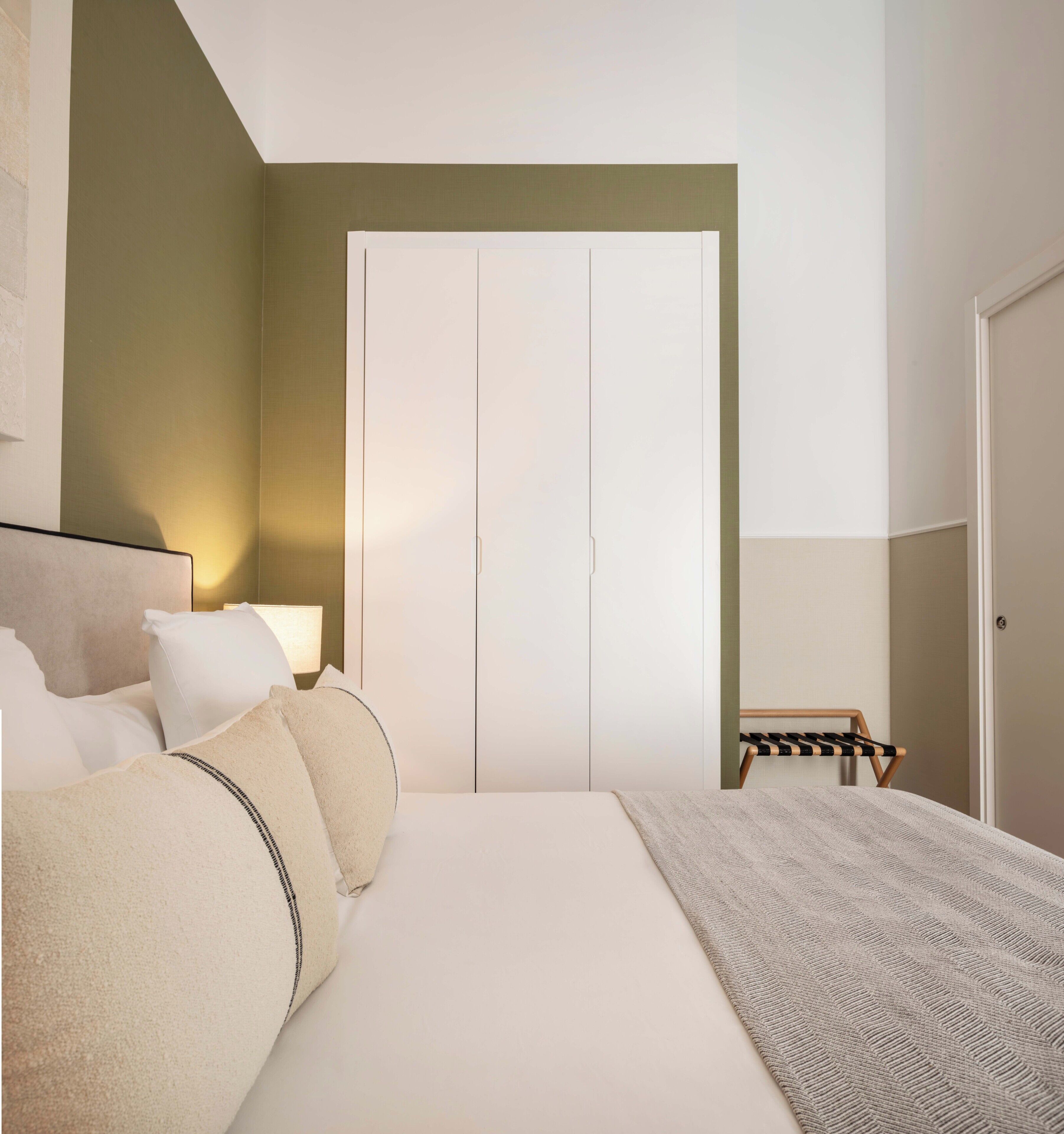 Un dormitorio con cama, almohadas, una mesita de noche con lámpara, una silla y un armario.
