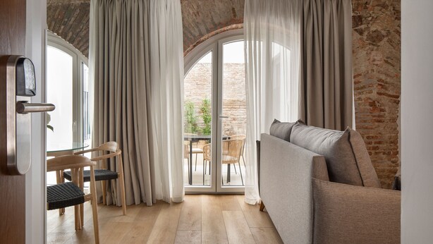 A room with a sofa, a dining table with chairs, and a view through a glass door to an outdoor area with a brick wall. - Feelathome Palacio de Álamos (Distrito Centro)