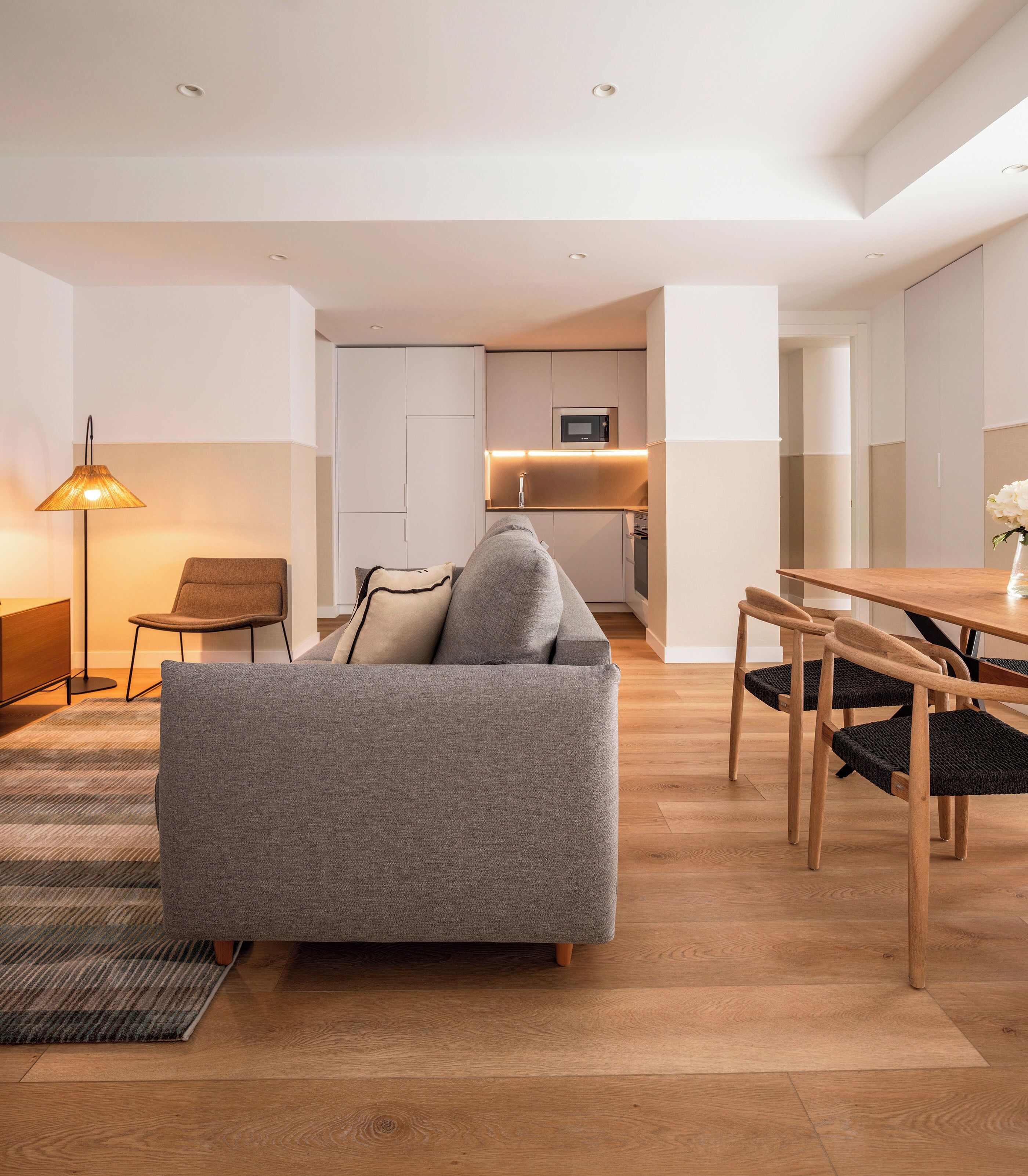 A modern living room with a grey sofa, a wooden dining table, and a kitchen in the background.
