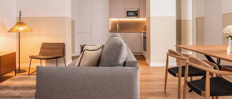 A modern living room with a grey sofa, a wooden dining table, and a kitchen in the background.