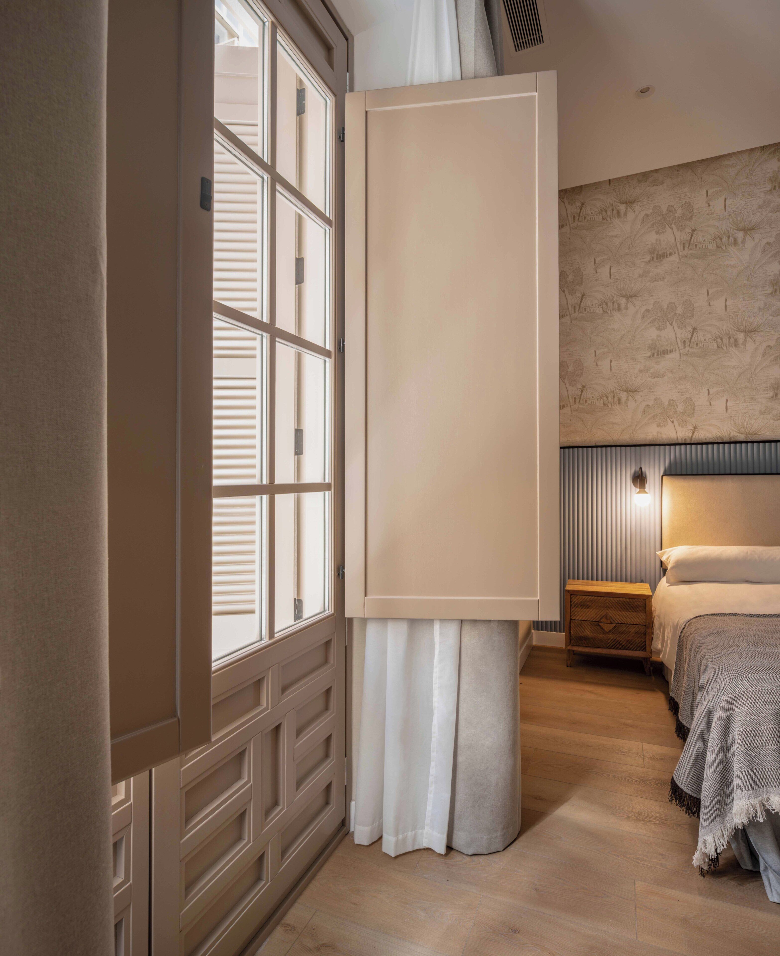 Une chambre avec une grande fenêtre, un lit doté d’une tête de lit, une table de chevet et un mur orné d’un motif texturé.