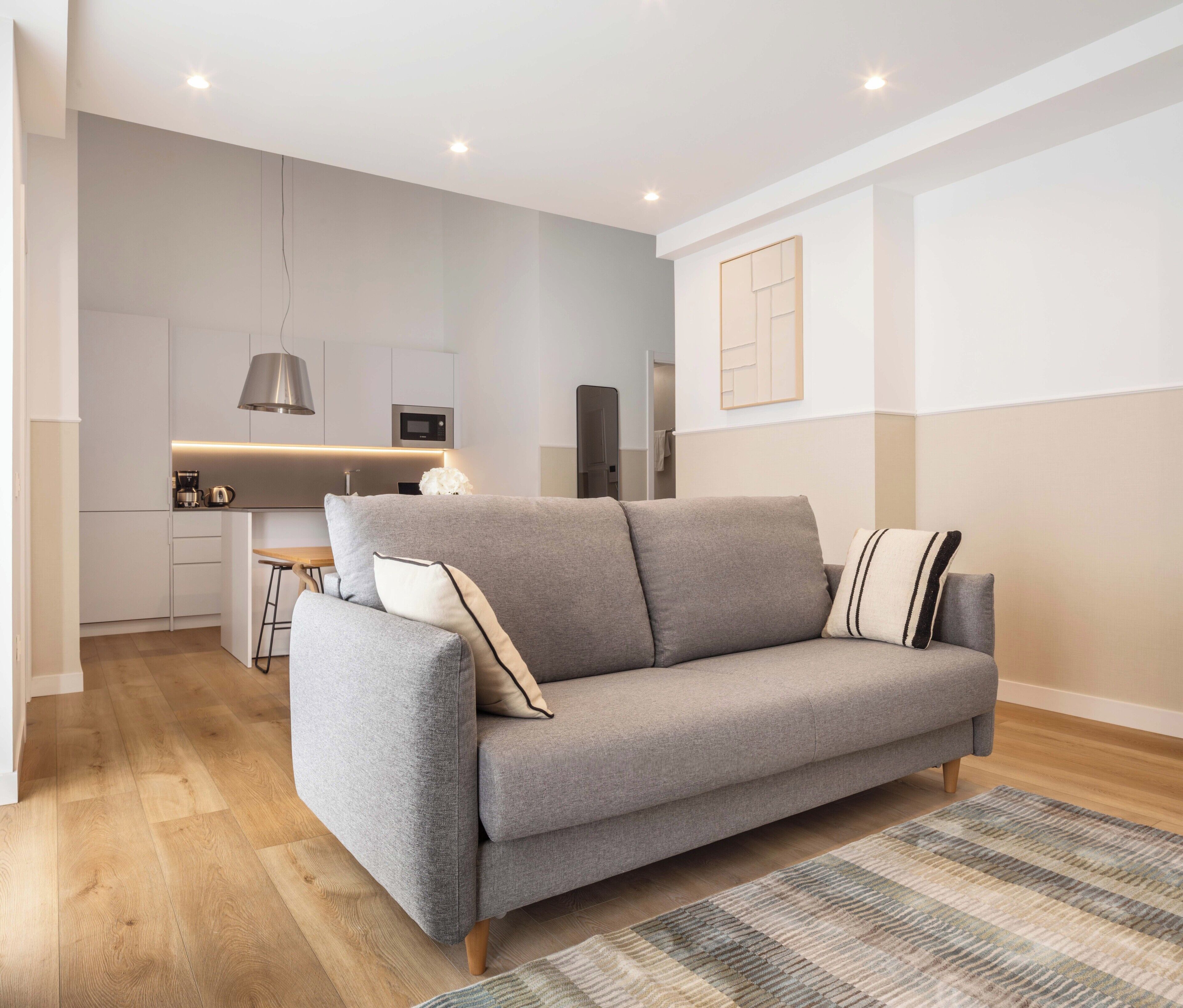 Un salon moderne avec un canapé gris, un plancher en bois et une cuisine en arrière-plan.