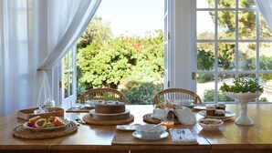 A breakfast table set with various dishes, including a cake, fruit, and a bowl of orange juice, with a view of greenery outside. - Luxury Oceanfront Villa - Catered & Serviced with Private Boat & Skipper (Tavira)