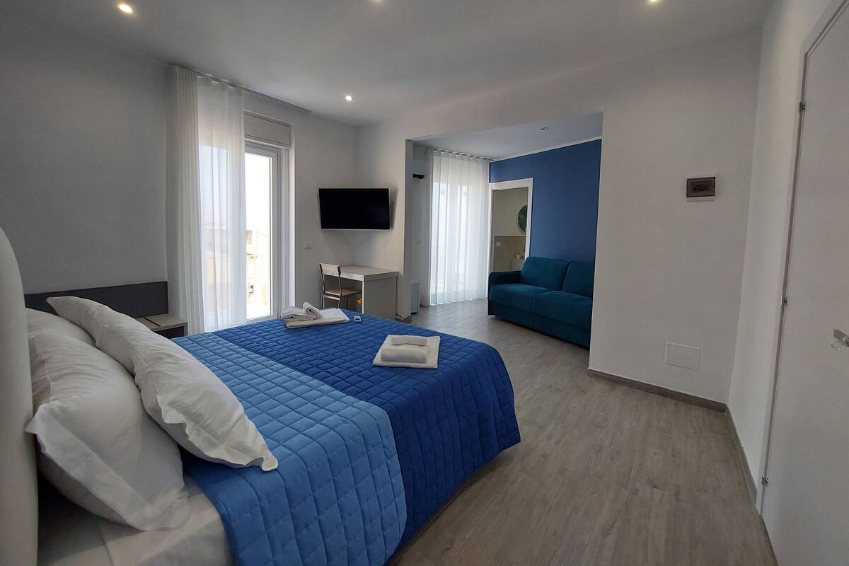 Una habitación de hotel moderna con una cama grande, un escritorio, un televisor de pantalla plana y un sofá en la zona de estar contigua.