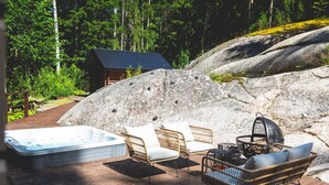 A wooden deck with a hot tub, wicker chairs, and a grill area, surrounded by large rocks and a cabin in a forest. - Stay North - Västerskog (Sipoo)