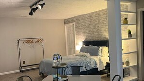 A bedroom with a bed, a desk, chairs, a hanging clothes rack, and a white brick wall. - Private Cozy Studio (Radford)