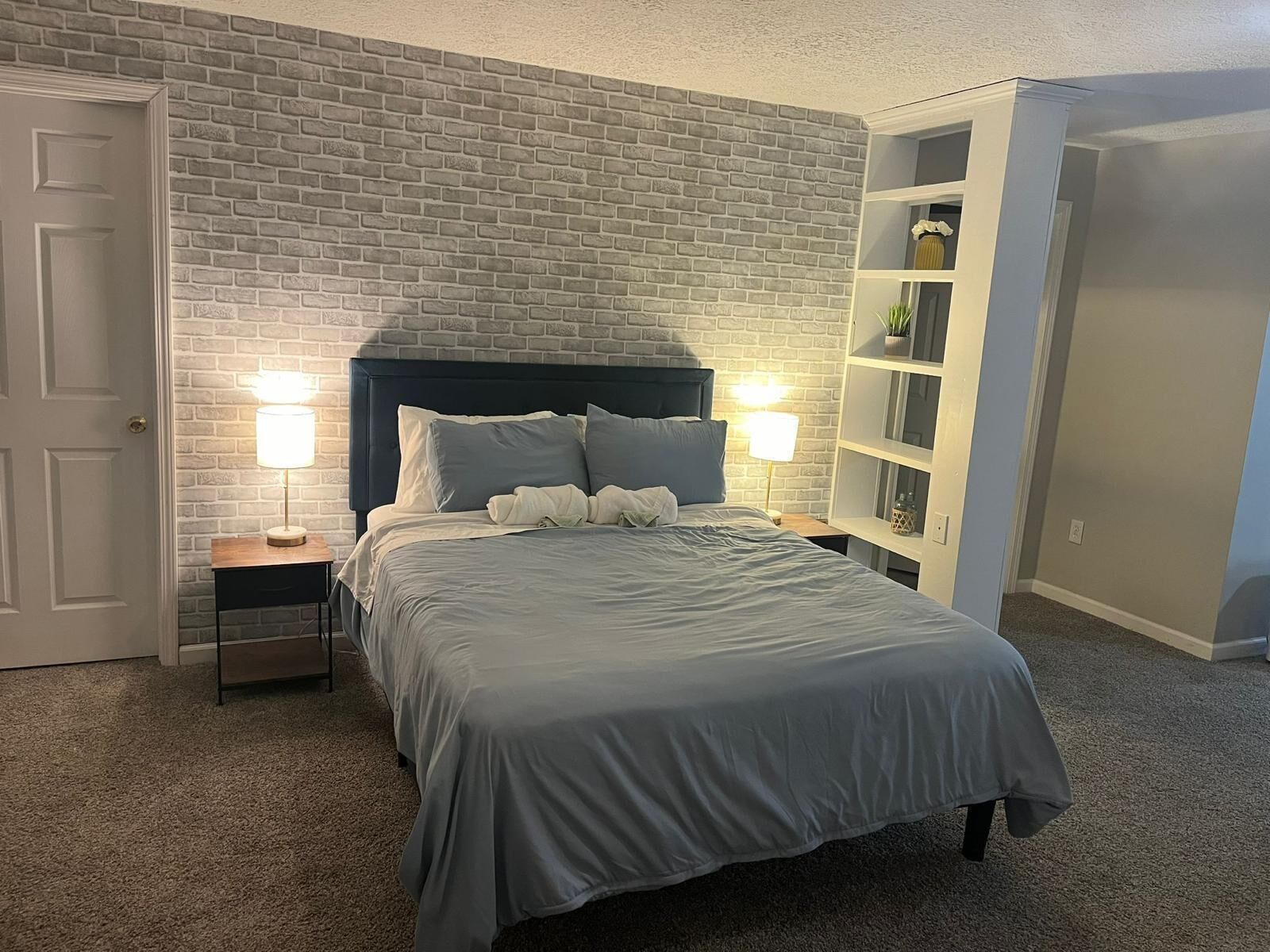 Un dormitorio con pared de ladrillo, una cama con cabecera oscura, mesitas de noche con lámparas y una estantería blanca.