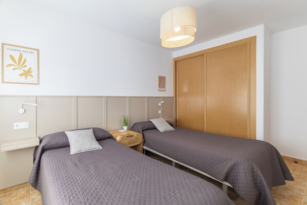 A bedroom with two beds, a wooden wardrobe, a bedside table with a plant, and a wall-mounted light fixture.
