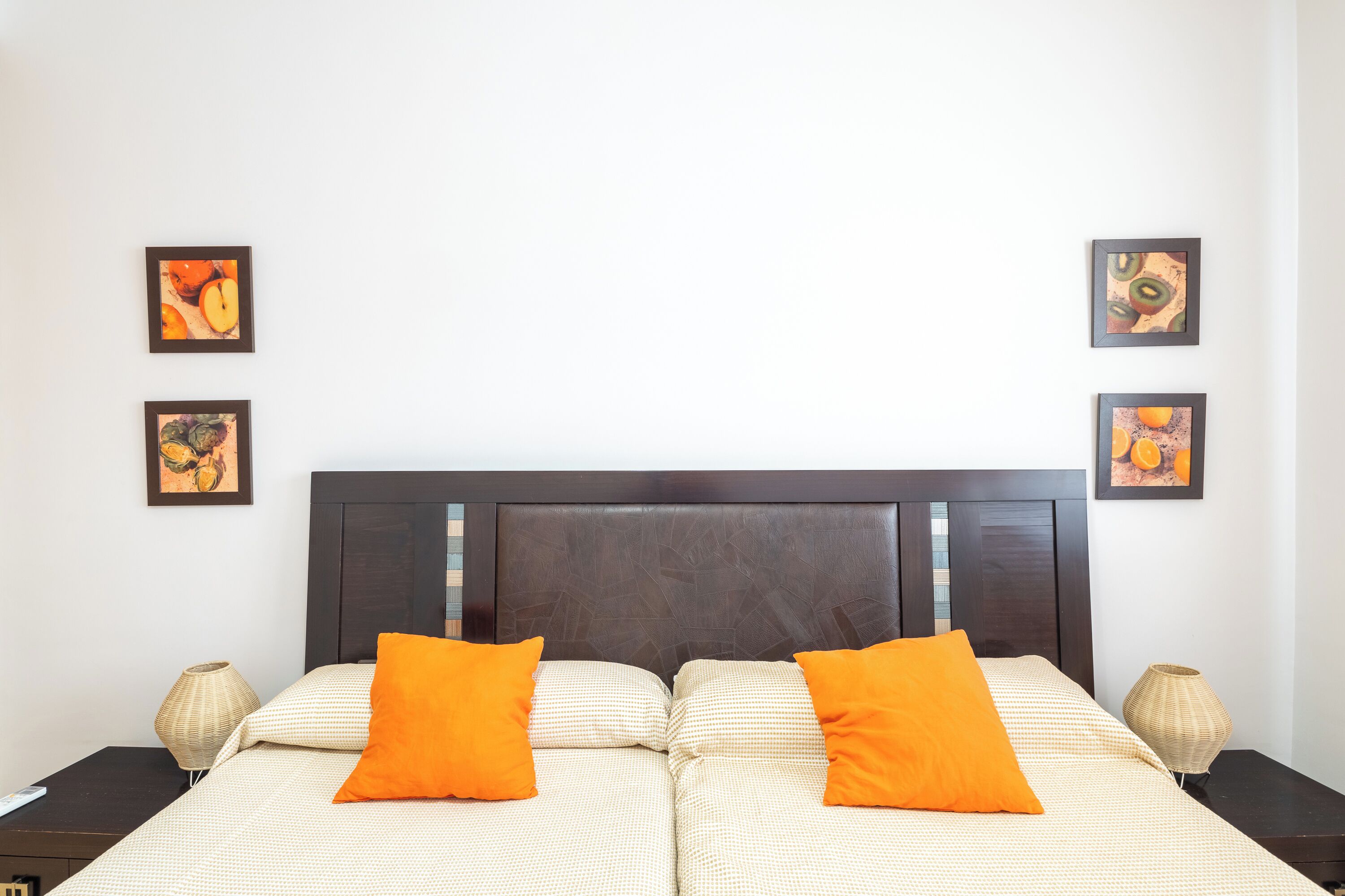 Ein Doppelbett mit zwei orangefarbenen Kissen, ein dunkles Kopfstück und vier gerahmte Kunstwerke an der Wand.