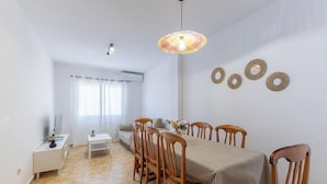 Un coin repas avec une table dressée pour six personnes, entourée de chaises en bois. Une console blanche, sur laquelle se trouvent un téléviseur et une petite plante, est adossée au mur.