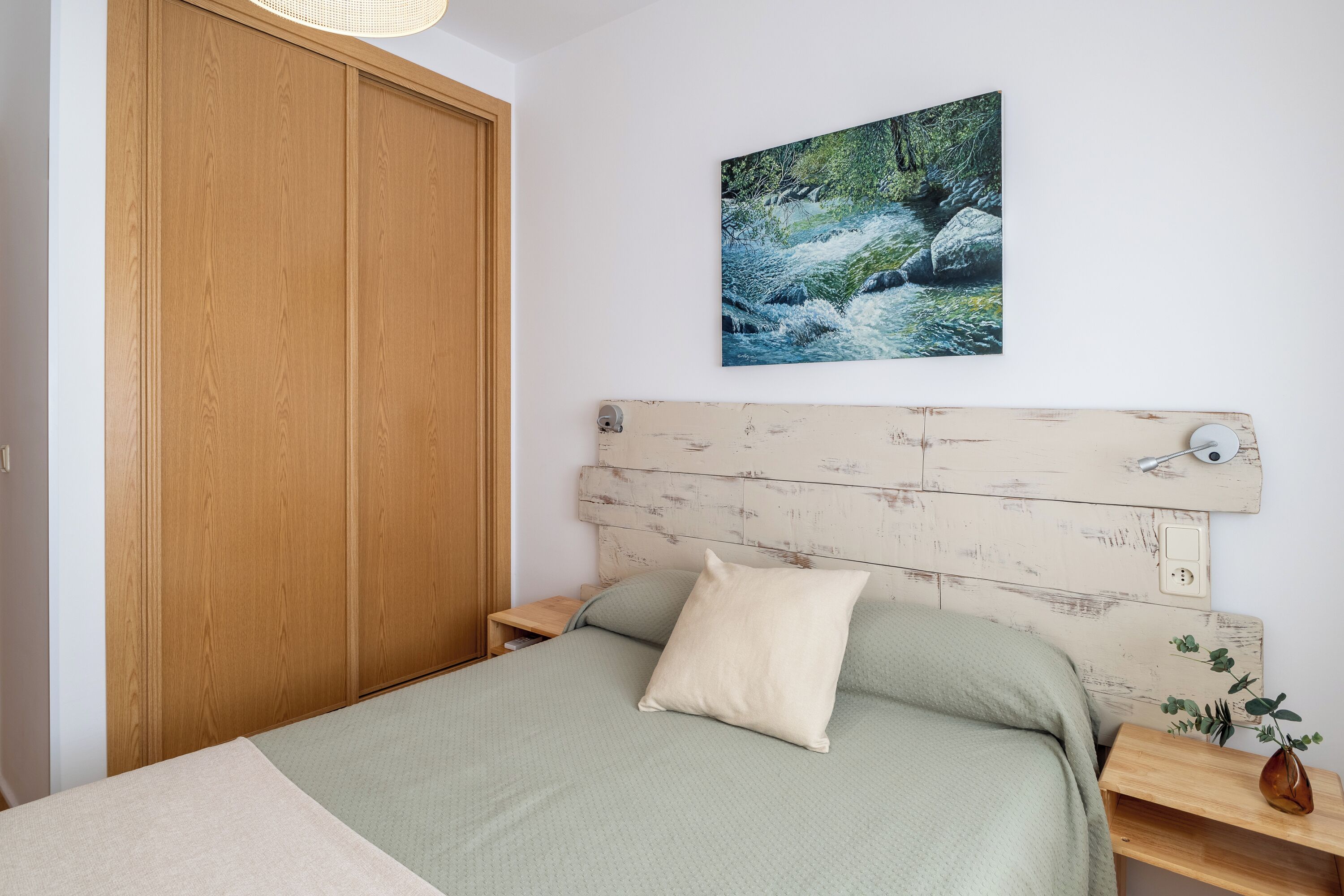 Une chambre avec une armoire en bois, un lit recouvert d’une couverture verte, un oreiller blanc, une table de chevet en bois ornée d’un vase, et un tableau représentant une rivière accroché au mur.
