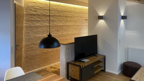 Una sala de estar moderna con pared de madera, una lámpara colgante negra, un televisor de pantalla plana sobre un soporte de madera y un pequeño otoman.