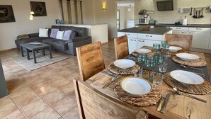 A modern living room with a grey sofa, a wooden dining table set for four, and a kitchen in the background. - Has und Igel - Refuge in Cala Santanyi with Private Pool and Barbecue (Cala Santanyí)