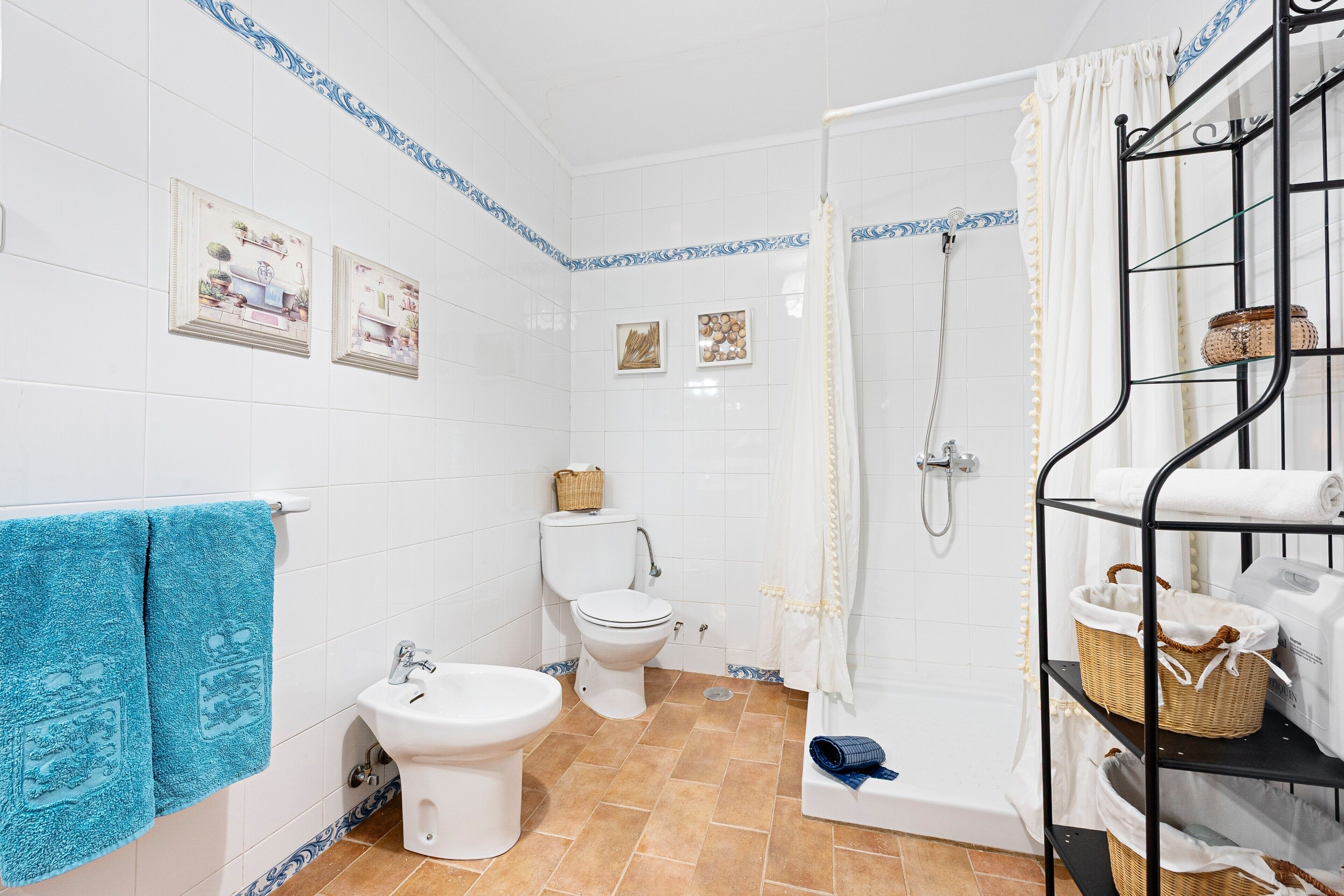 Baño con azulejos blancos, inodoro, bidé, ducha y repisa con cestas.