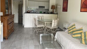 A compact living space with a kitchenette, a dining area with a glass table and metal chairs, a bed with striped pillows, and a wooden dresser. - 50 m from the beach - Beautiful garden apartment in a charming building (Antibes)