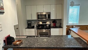 A kitchen with white cabinets, a granite countertop, a stainless steel stove, a microwave, and two white bar stools. - Charming 2-bedroom Stratton Mountain Condo with shuttle service (Stratton)
