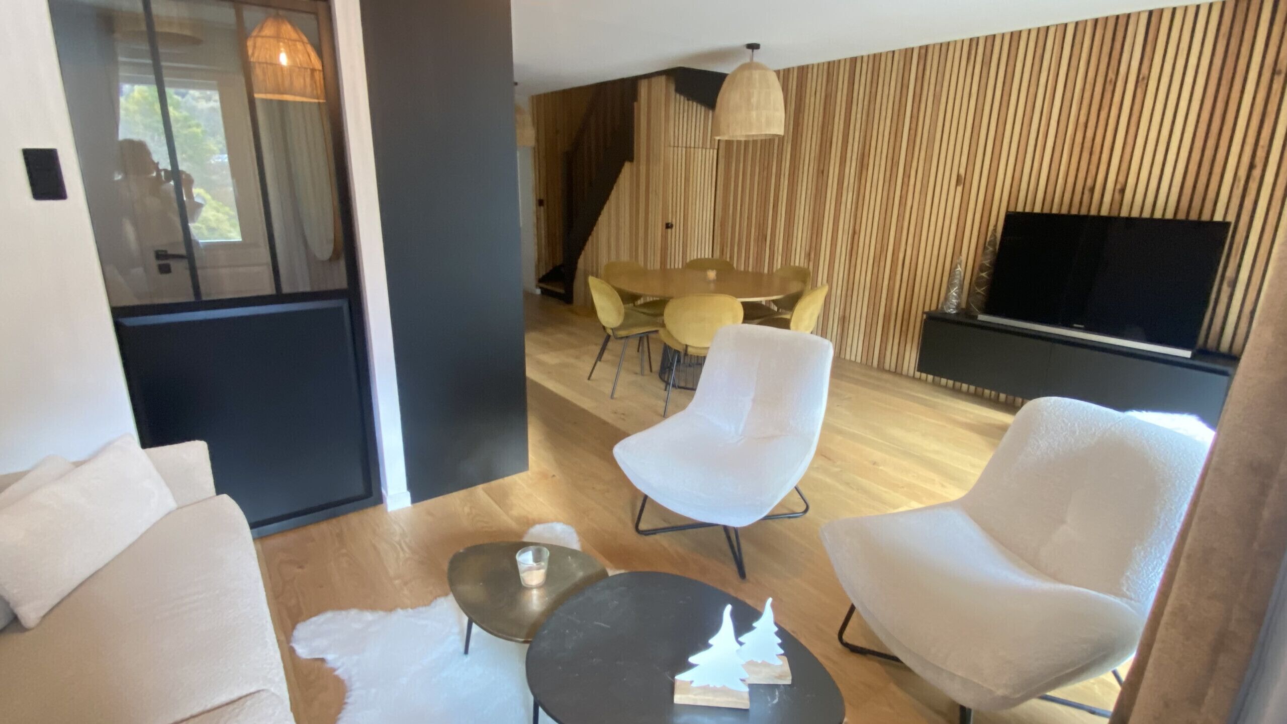 A modern living room with a sofa, two armchairs, a coffee table, and a dining area with a table and chairs. A flat-screen TV is mounted on the wall.